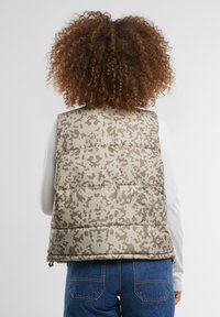 Gepolsterte Weste in Hellbraun mit Camouflage-Muster. Verfügt über einen Reißverschluss und ein gestuftes, gestepptes Design. Getragen über einem weißen Oberteil.