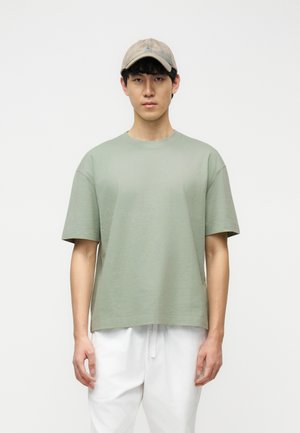 Noor mees, kes kannab beežikat nokamütsi, lahtist helerohelist lühikese varrukaga särki ja valgeid pükse, seisab tavalise valge tausta ees.