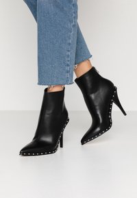 Mustat nahka nilkkurit, joissa on terävät varpaat, stiletto-korkokengät ja hopeiset niitit reunoilla. Yhdistetty lyhyisiin vaaleansinisiin farkkuihin.