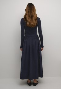 Marineblauwe jurk met lange mouwen, een nauwsluitend lijfje en een vloeiende rok, gemaakt van een gladde stof. De rok is geplooid bij de taille en wordt gedragen met platte schoenen.