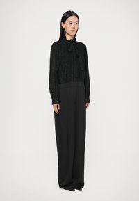 Blouse noire texturée avec un nœud papillon au col, associée à un pantalon noir à jambes larges. Les deux pièces ont une finition lisse et une silhouette ajustée.