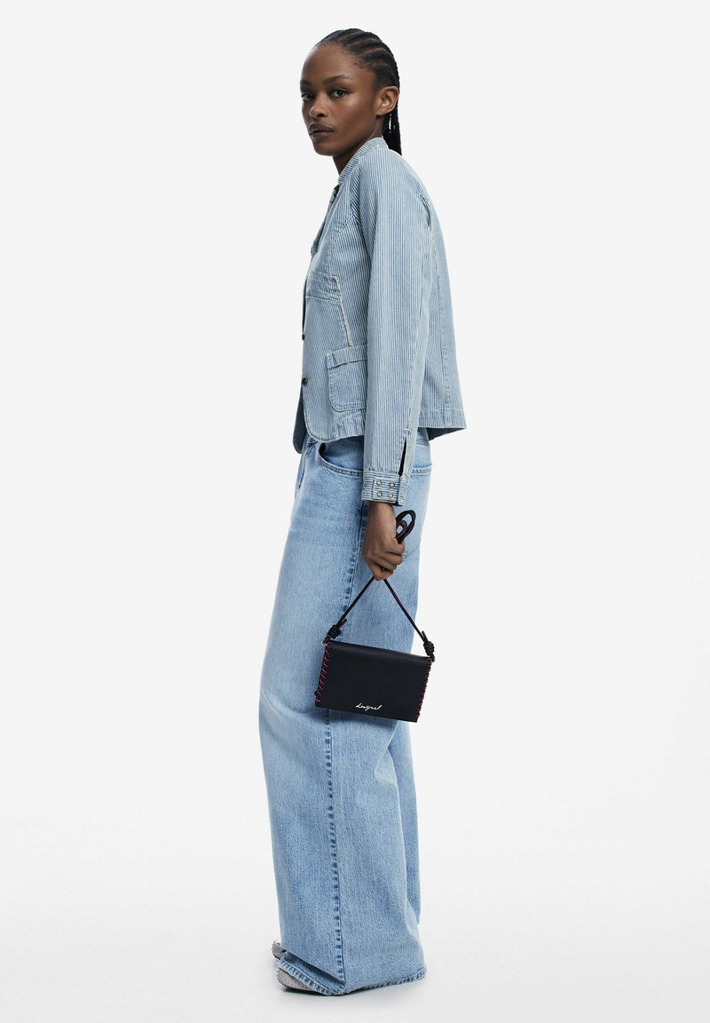 Hellblaue gestreifte Jeansjacke mit tailliertem Schnitt, kombiniert mit weit geschnittenen hellen Jeans. Hält eine kleine schwarze Handtasche mit roter Naht.