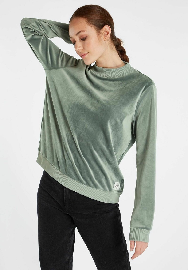 Fluwelen trui met lange mouwen in een gedempte groene kleur, met een hoge hals en een geribbelde zoom. Soepele textuur met een ontspannen pasvorm, gecombineerd met zwarte jeans.