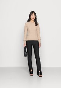 Top de mangas longas em bege às riscas combinado com calças pretas de bainha dividida. A modelo segura uma bolsa tecida preta e usa saltos abertos pretos.