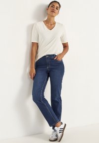 Ljust beige V-ringad tröja, blå denimjeans med synlig sömnad, och vita sneakers med svarta ränder och bruna sulor.