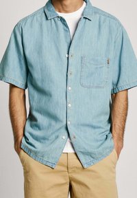 Camisa de mezclilla de manga corta azul claro con un bolsillo en el pecho, cierre de botones en la parte delantera y botones pálidos, llevada sobre una camiseta blanca. Tejido texturizado.