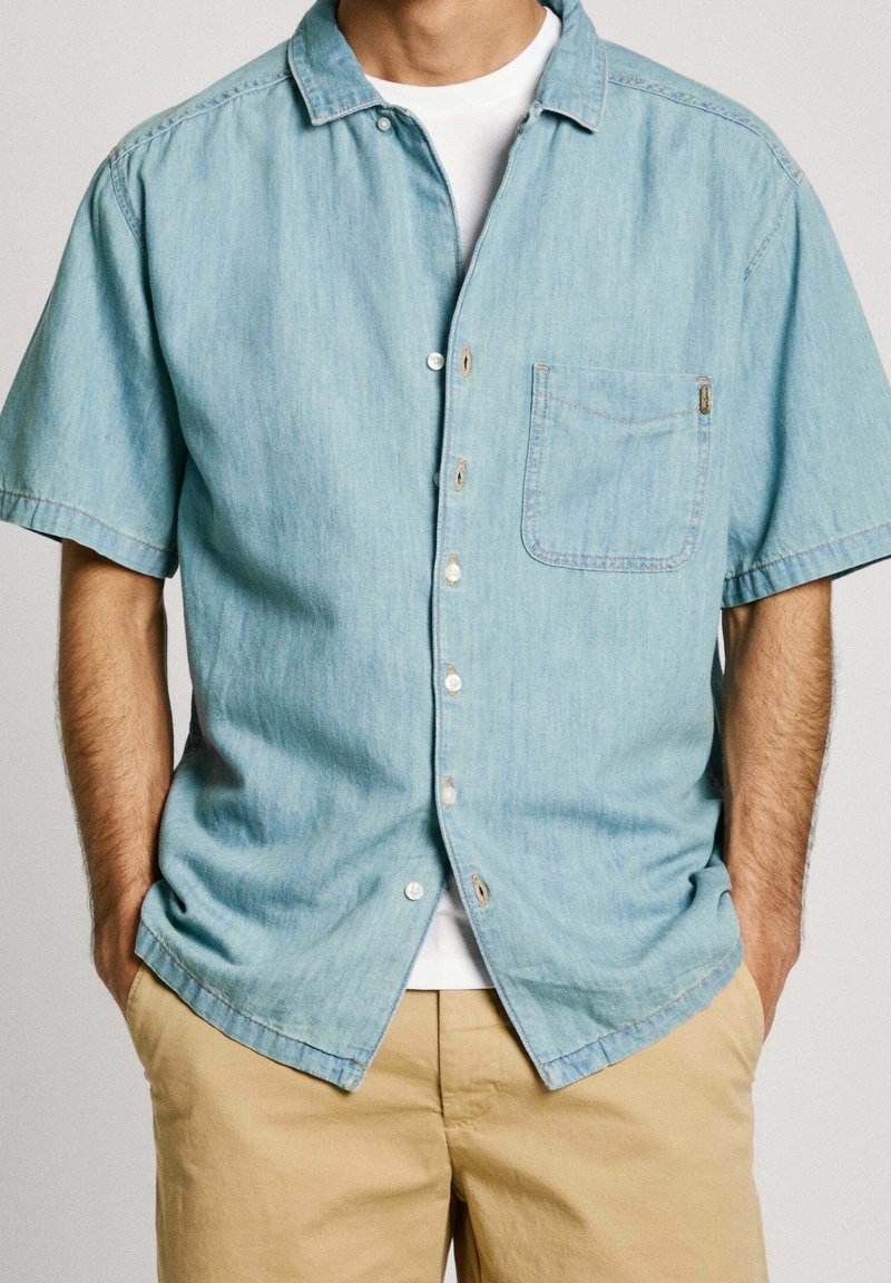 Camisa de mezclilla de manga corta azul claro con un bolsillo en el pecho, cierre de botones en la parte delantera y botones pálidos, llevada sobre una camiseta blanca. Tejido texturizado.