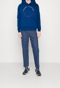 Námořnické modré mikina s kapucí a přední kapsou, s bílým a červeným kruhovým textem. V kombinaci s modrými džínami a tmavými teniskami.