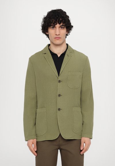 Olivově zelený bavlněný blazer s límečkem, třemi knoflíky, dvěma předními kapsami a texturovaným povrchem. Nosený přes černou košili.