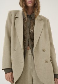 Beige dobbelknappet blazer med store knapper, tekstureret stof og skarpe revers, lagd over en grøn blondebluse med blomsterdetaljer.