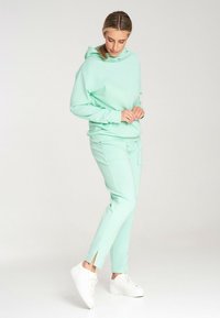 Miętowy kaptur i pasujące spodnie z gładkiego materiału. Kaptur ma sznurek do regulacji; spodnie mają boczne rozcięcia i kieszenie. Białe sneakersy dopełniają zestaw.