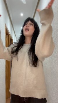 Νεαρή γυναίκα με μακριά σκούρα μαλλιά, φορώντας μπεζ πουλόβερ, δείχνει προς τα πάνω με τα δύο χέρια σε ένα διάδρομο με ξύλινες πόρτες.