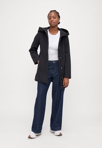 Mustad must jacket kapuutsiga, nööpidega ja küljetaskutega, koos laia säärega siniste teksade ja valgete jalanõudega.