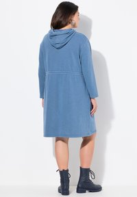 Rotes Kapuzenkleid aus weichem Stoff, mit langen Ärmeln, tailliertem Schnitt und seitlichen Taschen, getragen mit schwarzen Schnürstiefeletten.