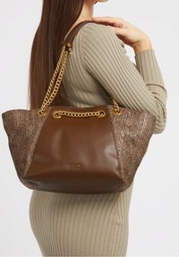 Borsa a mano in pelle marrone con dettagli intrecciati, manici a catena dorata e dettaglio del logo. Design spazioso, con parte superiore strutturata e texture morbida.