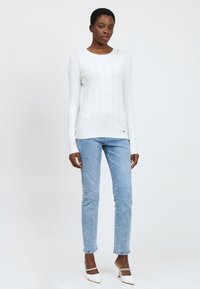 Suéter blanco de manga larga con un patrón texturizado, combinado con jeans de color azul claro. El modelo lleva zapatos de tacón blancos con una correa decorativa.