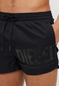 Fekete úszónadrág rugalmas derékrésszel és húzózsinórral. Jellemzője a tónusos "DIESEL" logó és a "SIKERES ÉLET EZER FORRÁSA" felirat.