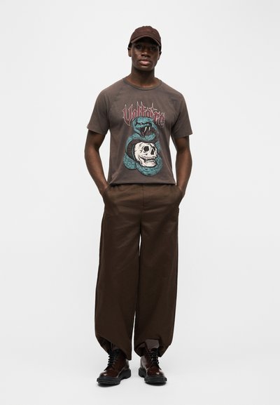 Άνδρας που φορά καφέ γραφικό t-shirt με σχέδιο φίδι και κρανίο, καφέ παντελόνι με φαρδιά μπατζάκια, σκούρα καφέ μπότες και καφέ καπέλο που ταιριάζει, στέκεται με τα χέρια στις τσέπες.