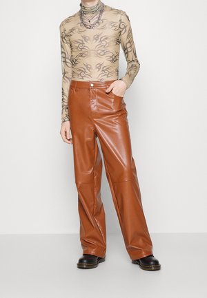 Top transparente de manga larga con patrones tribales, combinado con pantalones de pierna ancha de cuero sintético marrón y botas negras gruesas. Collar plateado en capas.