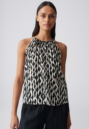 Femme portant un top sans manches à motif ondulé noir et blanc, associé à un pantalon noir, debout devant un fond gris uni.