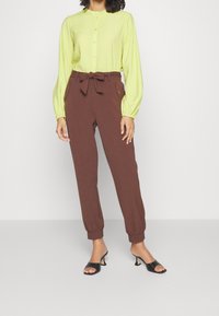 Kvinna som bär en limegrön långärmad blus med knappar, bruna högmidjade byxor med knytbälte och svarta sandaler med öppen tå.