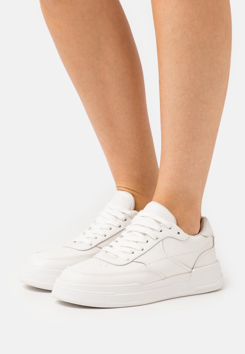 Person som bär vita låga sneakers med snörning, stående med fötterna nära varandra på en vit bakgrund.