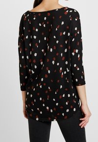 Schwarzes Oberteil mit 3/4-Ärmeln, das ein buntes gepunktetes Muster in Rot, Weiß und Beige aufweist und aus einem weichen Stoff gefertigt ist.