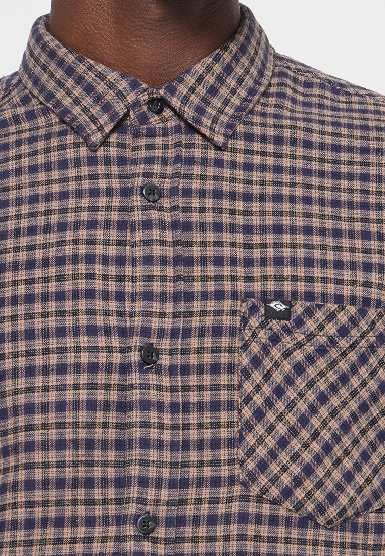 Gros plan sur une personne portant une chemise à carreaux bleu marine et beige avec une poche sur le devant et des boutons noirs.