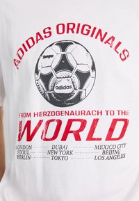 Hvit t-skjorte med et grafisk trykk av en svart-hvit fotball. Teksten sier "ADIDAS ORIGINALS" i rødt, med bynavn nederst.