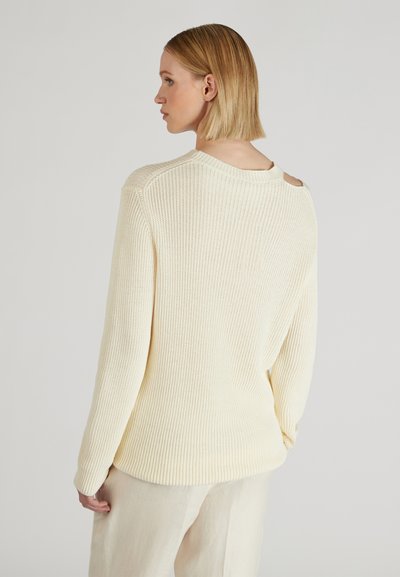 Donna con capelli biondi corti che indossa un maglione a coste crema con dettaglio a taglio sulla spalla, abbinato a pantaloni chiari.