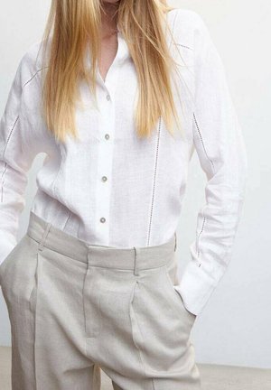 Osoba o długich blond włosach nosi białą koszulę na guziki, wpuszczoną w wysokowysokie beżowe plisowane spodnie, z rękami w kieszeniach.