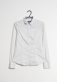 Vit skjorta med långa ärmar, med ett svart prickmönster och en liten broderad logotyp på bröstet. Hänger från en svart galge.