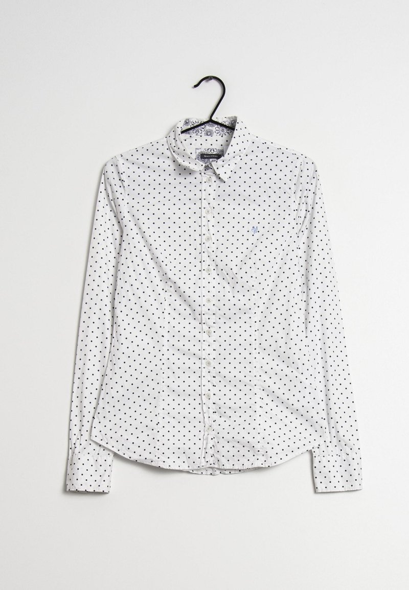 Vit skjorta med långa ärmar, med ett svart prickmönster och en liten broderad logotyp på bröstet. Hänger från en svart galge.