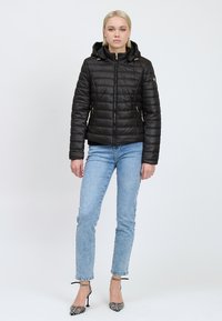 Sort pufferjakke med hætte og lynlåse i guldtoner, med et quiltet design, båret over blå jeans og mønstrede høje hæle.