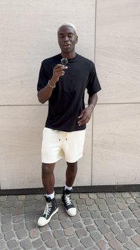 Svart kortärmad t-shirt, krämfärgade shorts och svarta högskurna sneakers med vita detaljer; står mot en ljus stenbakgrund.
