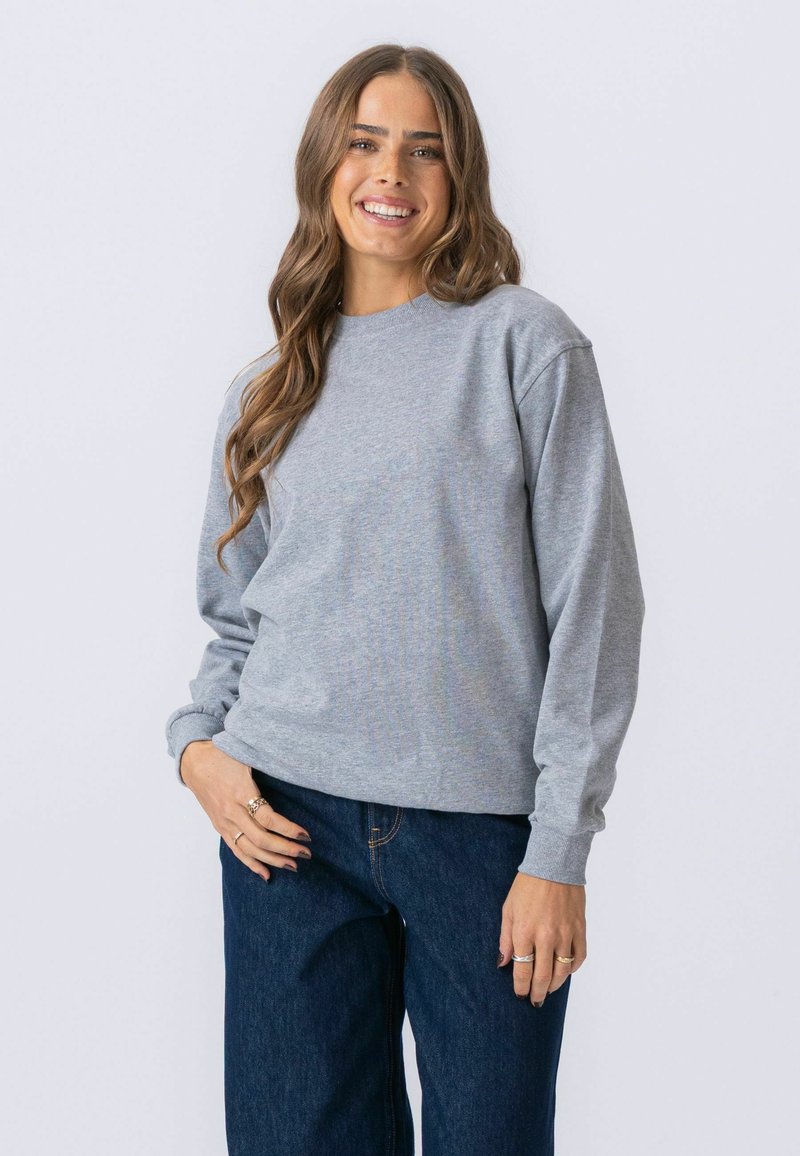 Jeune femme aux longs cheveux bruns portant un sweat-shirt gris clair et un jean bleu foncé, souriante avec une main dans la poche.