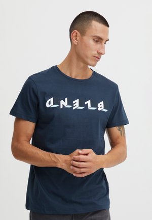 Moški s kratkimi lasmi, ki nosi temno modro majico z belim, stiliziranim besedilom "BLEND" napisanim od zadaj proti naprej, stoji pred enobarvnim svetlim ozadjem.