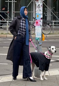 Kvinne i en mørk frakk og rutete skjorte med et blått skjerf står ved siden av en Dalmatiner i en svart skjorte, begge på fortauet i en by.