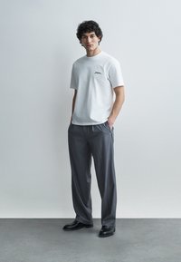 Moški z valovitimi lasmi stoji z rokami v žepih, oblečen v belo kratko majico s kratkimi rokavi, sive hlače in črne čevlje na enostavni podlagi.