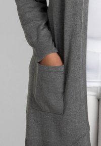 Grau gestrickte Strickjacke mit einer Fronttasche und gerippten Bündchen. Weiche Textur mit einem lockeren Schnitt, über einem weißen Oberteil getragen.
