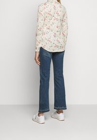 Blumenmuster langärmliges Hemd in Creme mit rosa und grünen Blumen, kombiniert mit blauen Schlagjeans und weißen Sneakers mit braunen Sohlen.