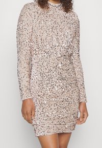 Kort klänning med lång ärm i pailletttyg i ljusrosa färg med rynkdetaljer som skapar en texturerad, skimrande effekt.