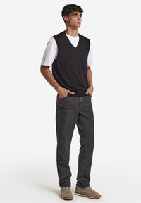 Mörkgrå ärmlös tröja över en vit t-shirt, kombinerad med mörka jeans och beiga skor; modellen står med händerna i fickorna.