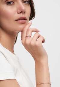 Roségoldring mit dem Wort "LIEBE" aus klaren Edelsteinen. Die Hand des Models liegt nah an ihrem Gesicht und trägt ein passendes Armband. Einfaches T-Shirt.
