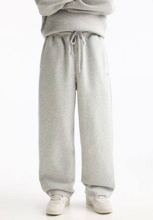 Personne debout portant un pantalon de jogging gris clair avec cordon à la taille et des baskets blanches sur fond blanc.