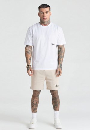 Mand med tatoveringer iført hvid t-shirt og beige korte bukser med matchende logoer, stående foran en ensartet lys baggrund.
