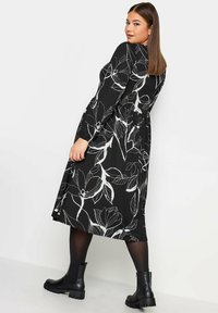 Musta mekko, jossa on valkoisia kukkakuoseja, pitkät hihat ja levenevä hame. Mustat nilkkurit, joissa on teksturoidut pohjat, täydentävät asua.