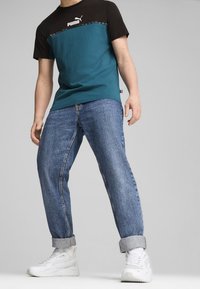 Homme portant un t-shirt Puma vert sarcelle et noir, un jean bleu retroussé et des baskets blanches, debout devant un fond gris uni.