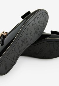 Zapatos de charol negros con un acento dorado y un detalle de lazo, que cuentan con una suela de goma texturizada grabada con patrones florales.