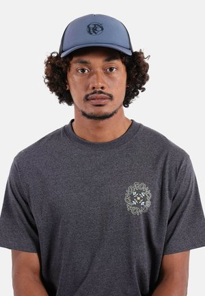 Homme aux cheveux bouclés portant une casquette de baseball bleue et un t-shirt gris foncé avec un graphique circulaire sur la poitrine gauche, faisant face vers l'avant.